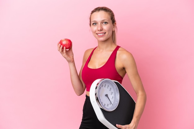 Jovem mulher caucasiana isolada em fundo rosa com máquina de pesagem e com uma maçã