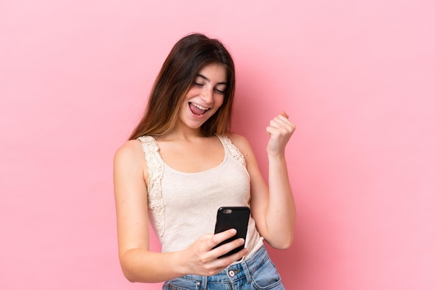 Jovem mulher caucasiana isolada em fundo rosa com telefone em posição de vitória