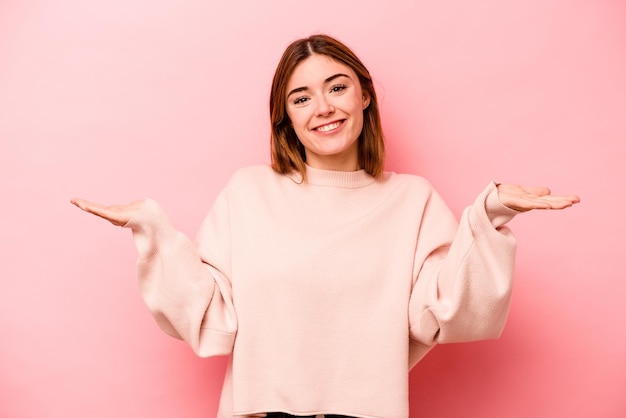 Jovem mulher caucasiana isolada em fundo rosa faz escala com braços se sente feliz e confiante