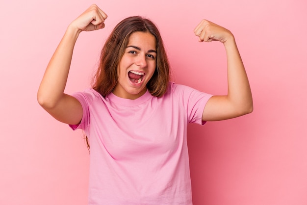 Jovem mulher caucasiana isolada em fundo rosa mostrando força gesto com os braços, símbolo do poder feminino