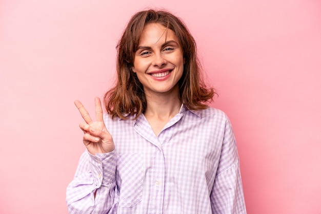 Jovem mulher caucasiana isolada em fundo rosa mostrando sinal de vitória e sorrindo amplamente