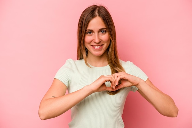 Jovem mulher caucasiana isolada em fundo rosa sorrindo e mostrando uma forma de coração com as mãos