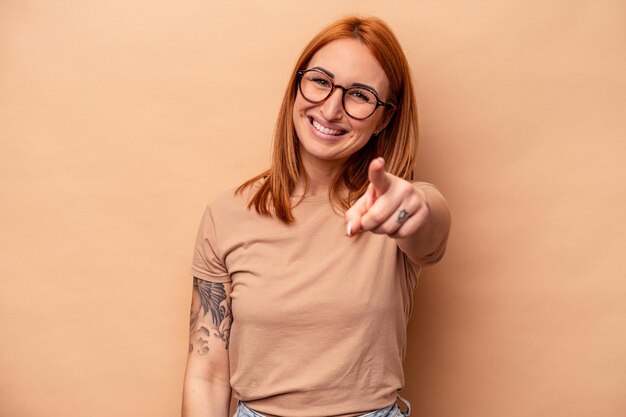 Jovem mulher caucasiana isolada em sorrisos alegres de fundo bege apontando para a frente.