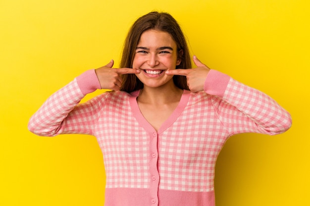 Foto jovem mulher caucasiana isolada em sorrisos de fundo amarelo, apontando o dedo para a boca.