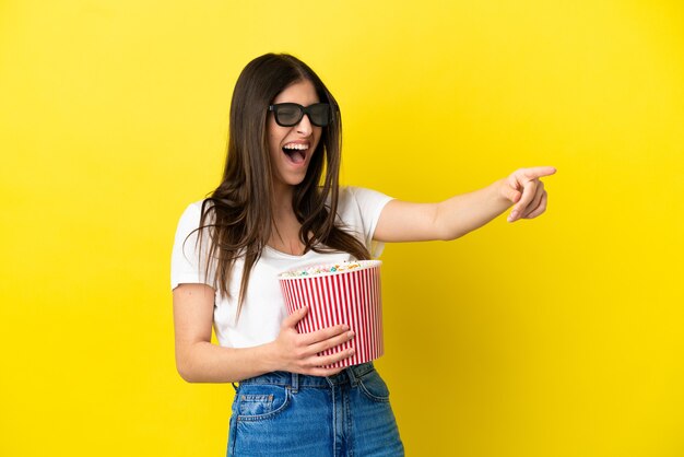 Jovem mulher caucasiana isolada em um fundo amarelo com óculos 3d e segurando um grande balde de pipocas enquanto aponta para longe