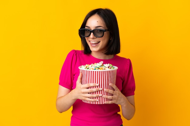 Foto jovem mulher caucasiana isolada em um fundo amarelo com óculos 3d e segurando um grande balde de pipocas