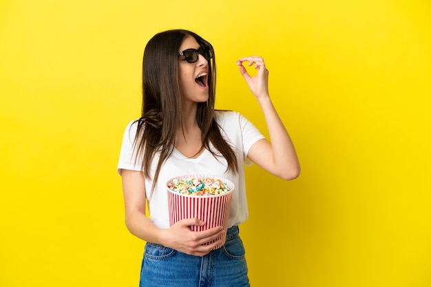 Jovem mulher caucasiana isolada em um fundo amarelo com óculos 3d e segurando um grande balde de pipocas