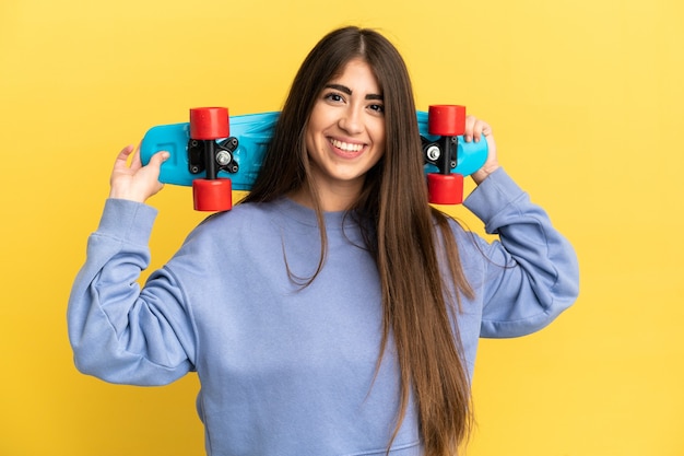 Jovem mulher caucasiana isolada em um fundo amarelo com um skate e uma expressão feliz