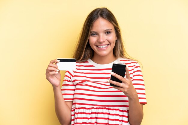 Jovem mulher caucasiana isolada em um fundo amarelo comprando com o celular com um cartão de crédito