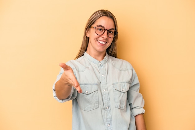 Foto jovem mulher caucasiana isolada em um fundo amarelo, esticando a mão na câmera em um gesto de saudação.