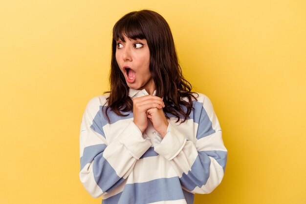 Jovem mulher caucasiana, isolada em um fundo amarelo, orando por sorte, espantada e abrindo a boca, olhando para a frente.