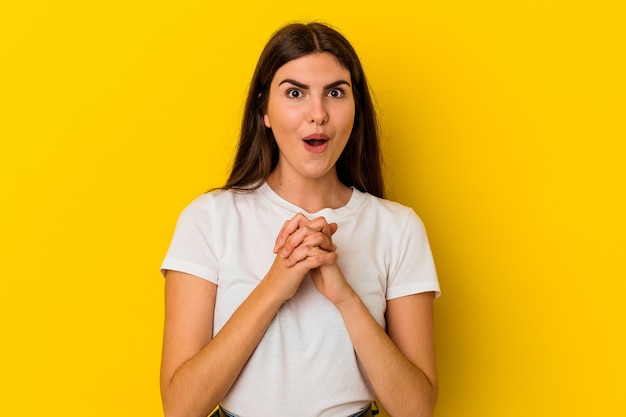 Jovem mulher caucasiana, isolada em um fundo amarelo, orando por sorte, espantada e abrindo a boca, olhando para a frente.