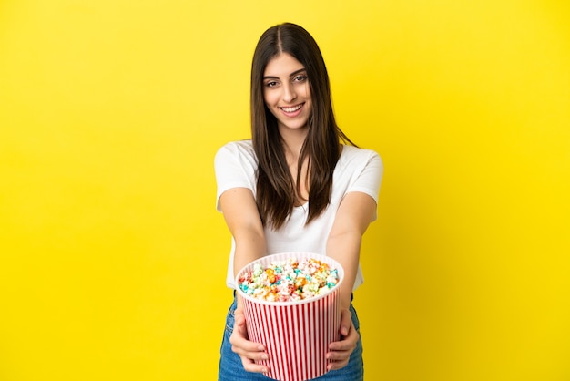 Jovem mulher caucasiana isolada em um fundo amarelo segurando um grande balde de pipocas