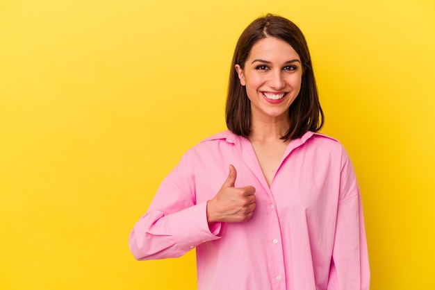 Jovem mulher caucasiana isolada em um fundo amarelo, sorrindo e levantando o polegar