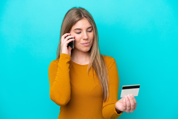 Jovem mulher caucasiana isolada em um fundo azul comprando com o celular com um cartão de crédito