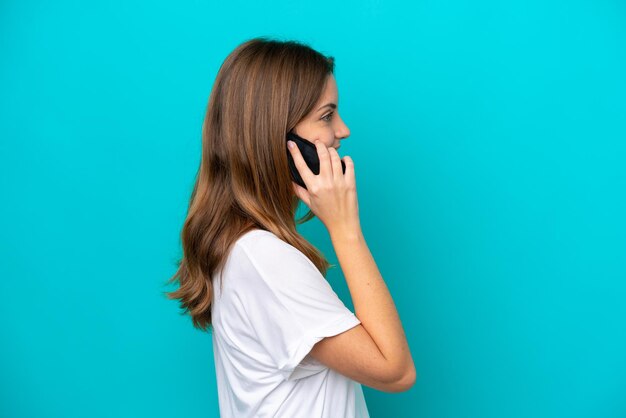 Jovem mulher caucasiana isolada em um fundo azul, conversando com alguém ao telefone celular