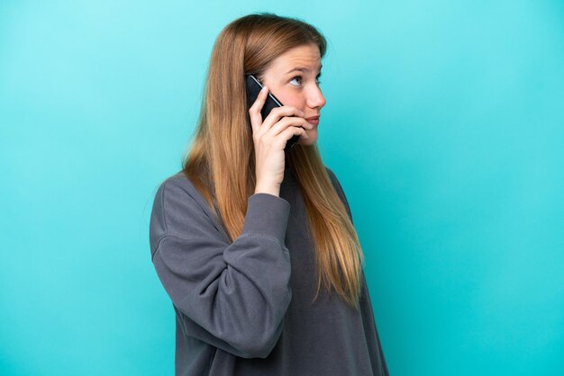 Jovem mulher caucasiana isolada em um fundo azul, conversando com alguém ao telefone celular