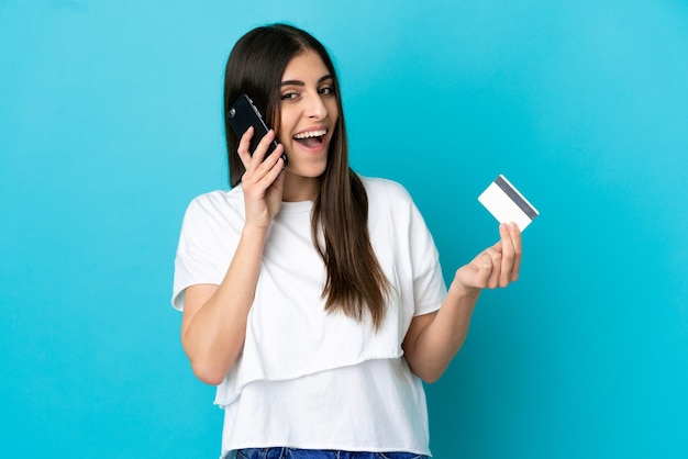 Jovem mulher caucasiana isolada em um fundo azul, conversando com o celular e segurando um cartão de crédito