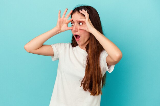 Jovem mulher caucasiana isolada em um fundo azul, mantendo os olhos abertos para encontrar uma oportunidade de sucesso.