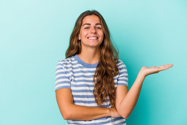 Jovem mulher caucasiana isolada em um fundo azul, mostrando um espaço de cópia na palma da mão e segurando a outra mão na cintura.