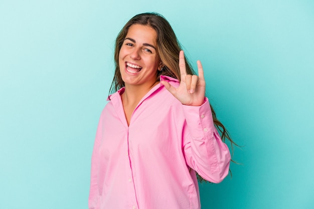 Jovem mulher caucasiana isolada em um fundo azul, mostrando um gesto de chifres como um conceito de revolução.