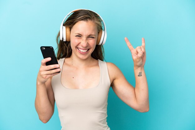 Jovem mulher caucasiana isolada em um fundo azul ouvindo música com um celular fazendo gesto de rock