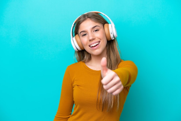 Jovem mulher caucasiana isolada em um fundo azul ouvindo música e com o polegar para cima