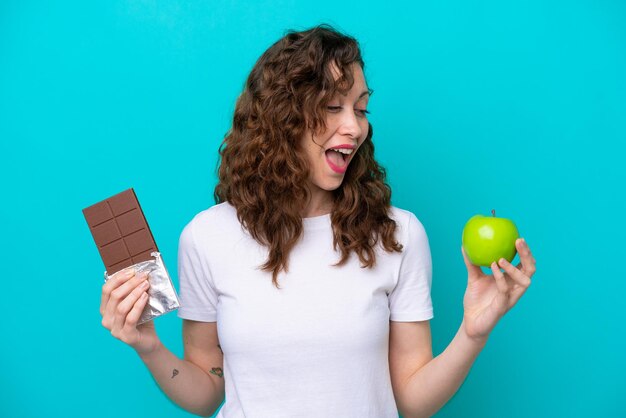 Jovem mulher caucasiana isolada em um fundo azul pegando um tablete de chocolate em uma mão e uma maçã na outra