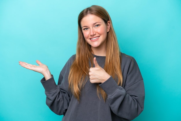 Jovem mulher caucasiana isolada em um fundo azul segurando copyspace imaginário na palma da mão para inserir um anúncio e com o polegar para cima