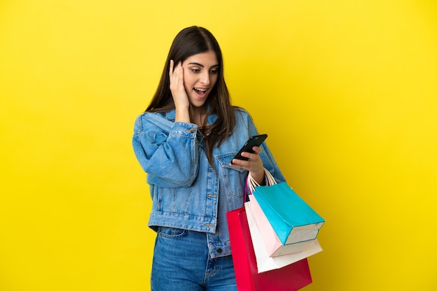 Jovem mulher caucasiana isolada em um fundo azul segurando sacolas de compras e escrevendo uma mensagem com seu celular para um amigo