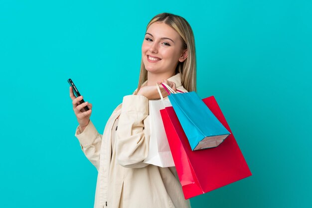 Jovem mulher caucasiana isolada em um fundo azul segurando sacolas de compras e escrevendo uma mensagem com seu celular para um amigo