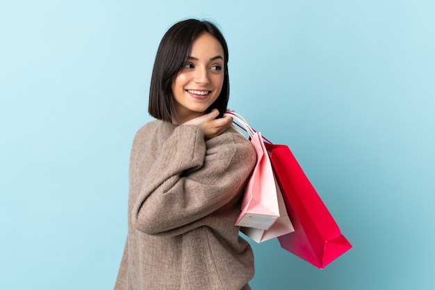 Jovem mulher caucasiana isolada em um fundo azul segurando sacolas de compras e olhando para trás