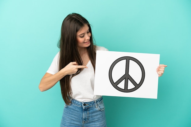 Jovem mulher caucasiana isolada em um fundo azul segurando um cartaz com o símbolo da paz e apontando-o