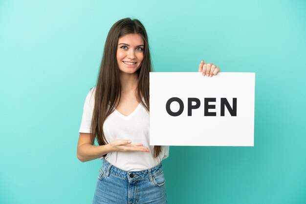 Jovem mulher caucasiana isolada em um fundo azul segurando um cartaz com o texto ABRIR e apontando-o