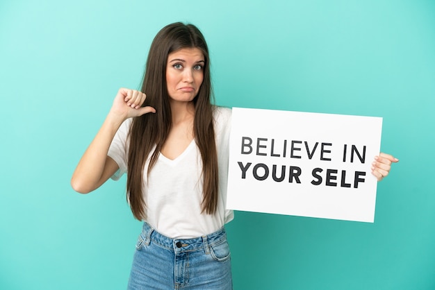 Foto jovem mulher caucasiana isolada em um fundo azul segurando um cartaz com o texto acredite em você mesmo com um gesto orgulhoso