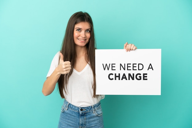 Jovem mulher caucasiana isolada em um fundo azul segurando um cartaz com o texto Precisamos de uma mudança com o polegar para cima