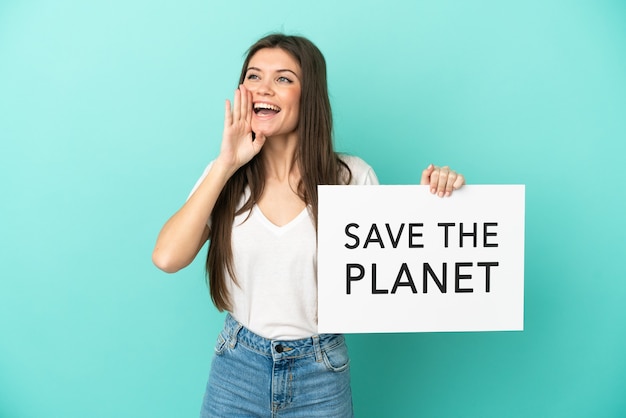 Jovem mulher caucasiana isolada em um fundo azul segurando um cartaz com o texto Salve o planeta e gritando