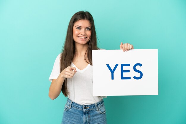 Jovem mulher caucasiana isolada em um fundo azul segurando um cartaz com o texto SIM e apontando-o