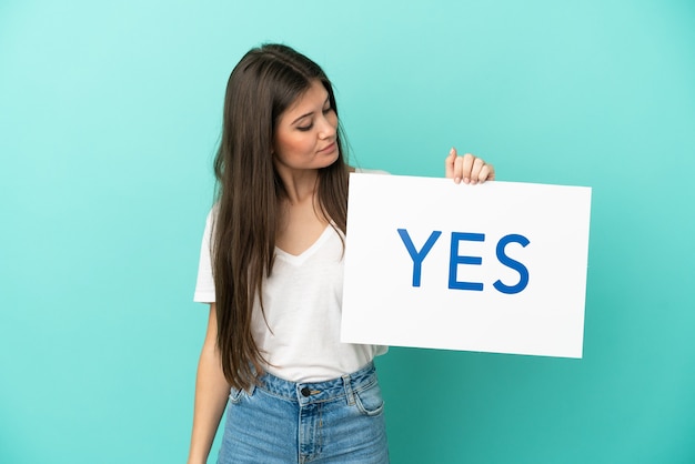 Jovem mulher caucasiana isolada em um fundo azul segurando um cartaz com o texto SIM