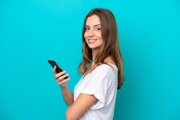 Jovem mulher caucasiana isolada em um fundo azul segurando um telefone celular e com os braços cruzados