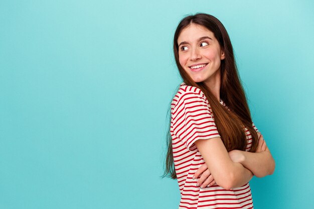 Jovem mulher caucasiana isolada em um fundo azul, sorrindo confiante com os braços cruzados.