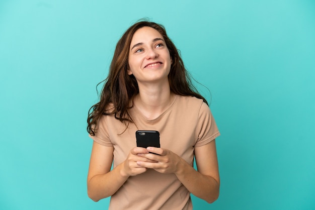 Jovem mulher caucasiana isolada em um fundo azul usando telefone celular e olhando para cima