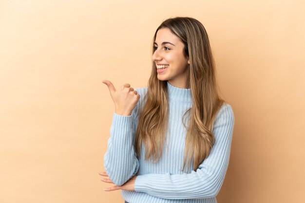 Jovem mulher caucasiana isolada em um fundo bege apontando para o lado para apresentar um produto