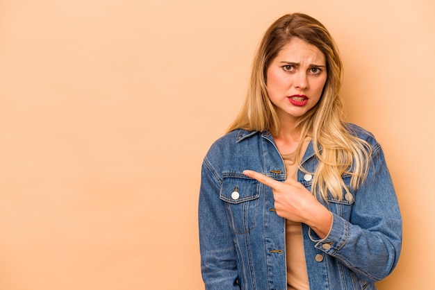 Jovem mulher caucasiana isolada em um fundo bege apontando para o lado