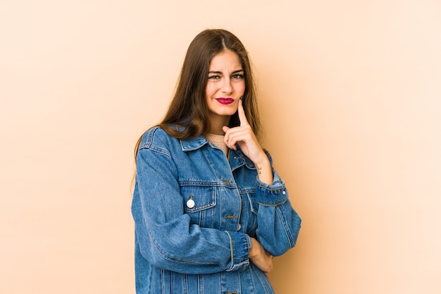 Jovem mulher caucasiana isolada em um fundo bege, contemplando, planejando uma estratégia, pensando sobre o funcionamento de um negócio.