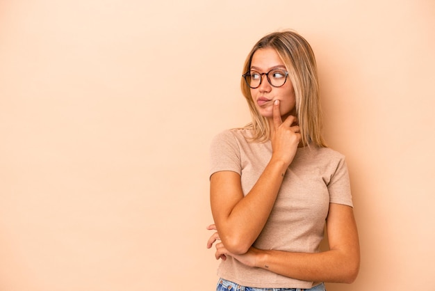 Jovem mulher caucasiana isolada em um fundo bege, olhando de soslaio com expressão duvidosa e cética.