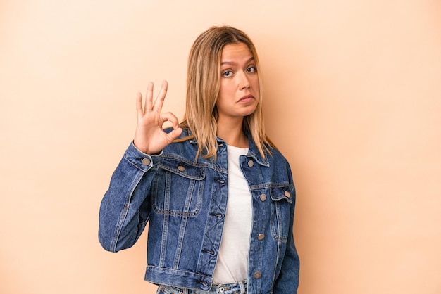 Jovem mulher caucasiana isolada em um fundo bege pisca um olho e segura um gesto de ok com a mão.