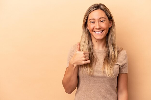 Jovem mulher caucasiana isolada em um fundo bege sorrindo e levantando o polegar