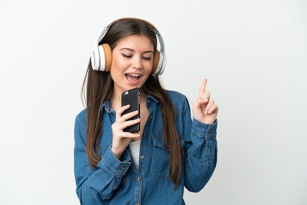 Jovem mulher caucasiana isolada em um fundo branco ouvindo música com um celular e cantando