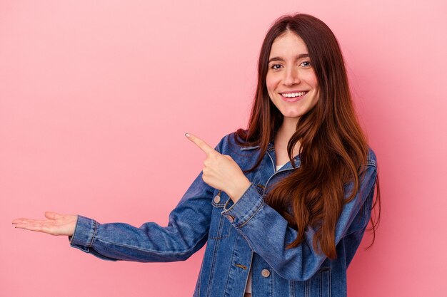 Jovem mulher caucasiana isolada em um fundo rosa animado segurando um espaço de cópia na palma da mão.